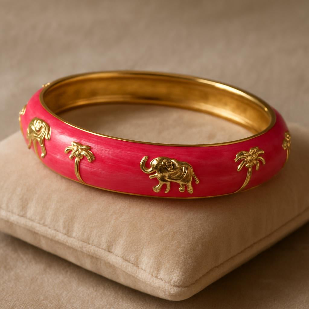 A close-up shot of the Pink Enamel Elephant Motif Bangle resting on a soft velvet cushion, highlighting the glossy pink enamel finish and intricate golden elephant and palm tree motifs, with gentle lighting emphasizing the shiny surface.