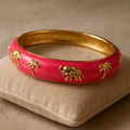 A close-up shot of the Pink Enamel Elephant Motif Bangle resting on a soft velvet cushion, highlighting the glossy pink enamel finish and intricate golden elephant and palm tree motifs, with gentle lighting emphasizing the shiny surface.