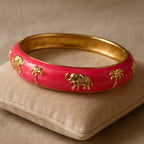A close-up shot of the Pink Enamel Elephant Motif Bangle resting on a soft velvet cushion, highlighting the glossy pink enamel finish and intricate golden elephant and palm tree motifs, with gentle lighting emphasizing the shiny surface.