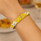 A close-up of the Sunny Yellow Floral Enamel Hinged Bangle on a woman's wrist, highlighting the glossy enamel flowers and sparkling crystal centers, with a blurred background of a brunch setting.